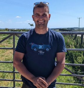 Thomas Marsh, co-owner of Eweleaze Dairy, smiling at a farm gate. He's wearing a dark t-shirt with a Land Rover graphic and has sunglasses pushed up on his head. Rolling green Dorset countryside and farm buildings visible behind him