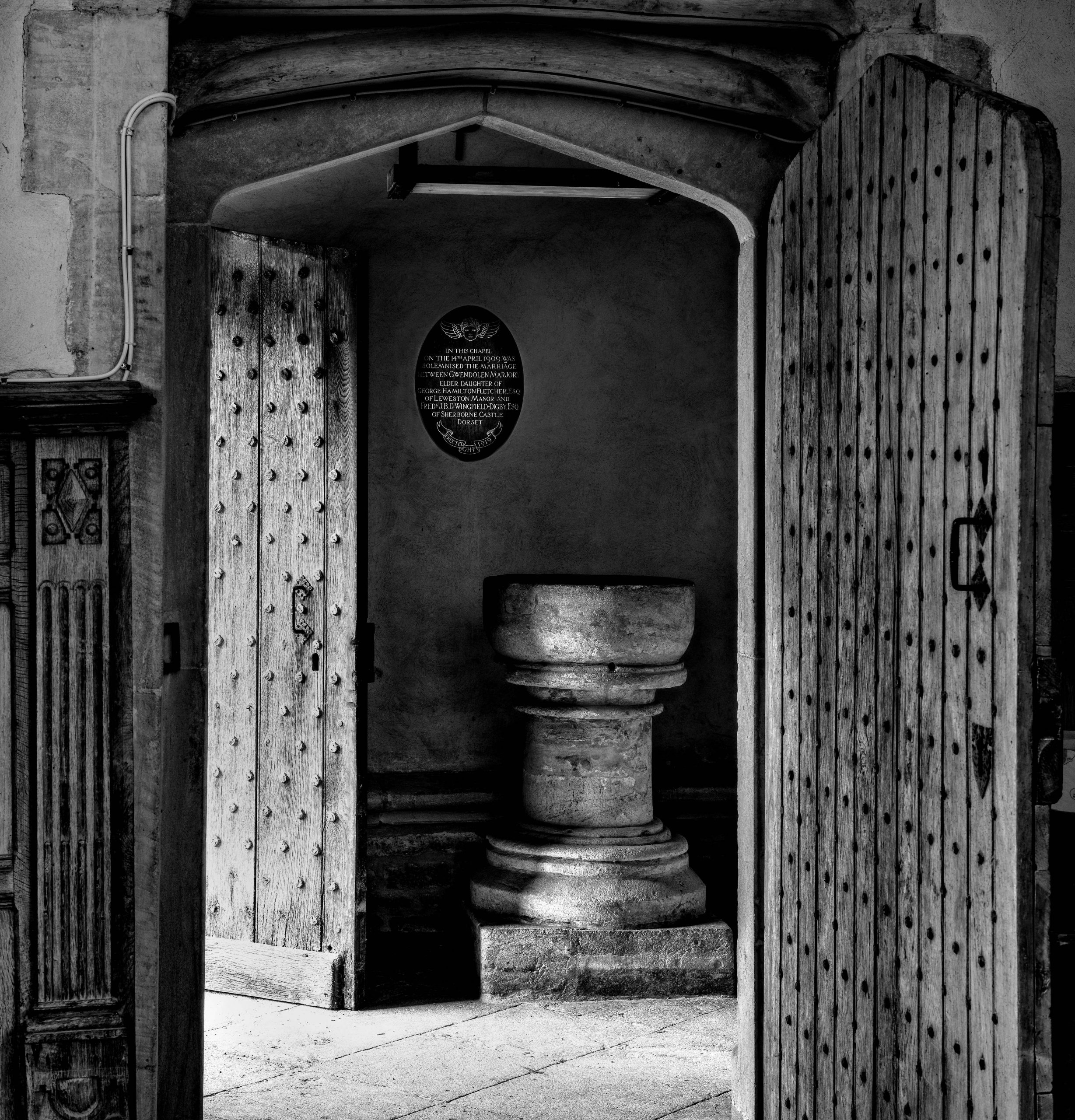 Black and white photograph of a stone baptismal font framed through heavy wooden doors studded with iron nails, showing the architectural detail and composition of Jim Johnson's church interior photography project"