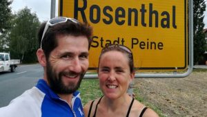 Justin, with dark hair and beard wearing a blue and white athletic jacket, and Amber, with dark hair in a black sports top, both smiling at the camera in front of a yellow Rosenthal Stadt Peine town sign