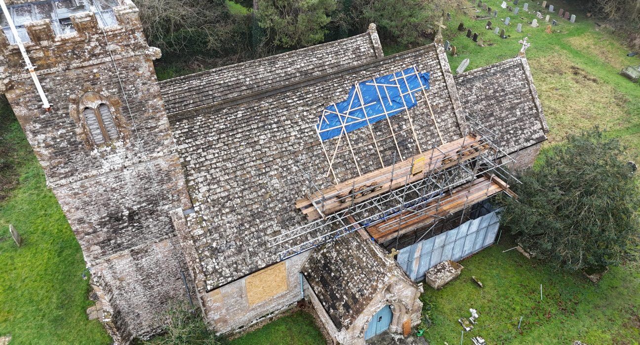 Aerial drone photograph of St Juthware Church in Halstock, showing the damaged roof from above. The church is built of weathered grey and honey-coloured stone, with a sturdy 15th-century tower on the left topped with battlements and a flagpole. A large section of the main roof's stone tiles has been covered with a bright blue tarpaulin, held down by a criss-cross frame of wooden battens, marking where rainwater has been getting in. Metal scaffolding with wooden walkways has been erected along the right side of the building, with stacks of tiles resting on the platforms. A small porch with a pale blue door sits at the front of the church, and one window below the roofline has been boarded over. The church is surrounded by green grass, with rows of headstones visible in the churchyard at the top right, and trees and hedgerows enclosing the scene. The image captures both the beauty of the historic building and the scale of the repair work needed.