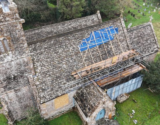 Aerial drone photograph of St Juthware Church in Halstock, showing the damaged roof from above. The church is built of weathered grey and honey-coloured stone, with a sturdy 15th-century tower on the left topped with battlements and a flagpole. A large section of the main roof's stone tiles has been covered with a bright blue tarpaulin, held down by a criss-cross frame of wooden battens, marking where rainwater has been getting in. Metal scaffolding with wooden walkways has been erected along the right side of the building, with stacks of tiles resting on the platforms. A small porch with a pale blue door sits at the front of the church, and one window below the roofline has been boarded over. The church is surrounded by green grass, with rows of headstones visible in the churchyard at the top right, and trees and hedgerows enclosing the scene. The image captures both the beauty of the historic building and the scale of the repair work needed.