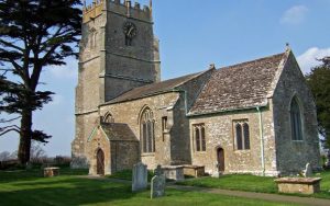 St Thomas à Becket Church, Lydlinch, a stone-built medieval church with a square tower, viewed from the churchyard. The church sits in a green landscape with gravestones in the foreground and a large cedar tree to the left. Clear blue sky and grass suggest a spring or summer day