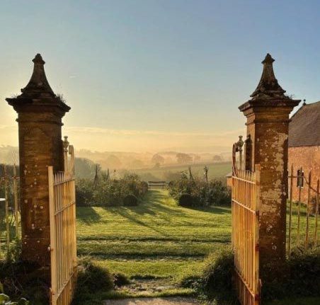 Alt text for Bettiscombe: "Looking through twin brick gateposts with decorative stone tops, framing a wooden gate, the view opens onto the sweeping gardens of Bettiscombe Manor. Rolling green lawns stretch into the distance, lined with mature trees and flowering borders, with the Dorset countryside and Vale beyond. The early morning or late afternoon light casts long shadows across the grass, and a soft mist hangs gently over the distant landscape beneath a pale blue sky. This is the entrance to designer Jasper Conran's Bettiscombe Manor near Bridport, which opens for the National Garden Scheme on Thursday 11 June. The gardens showcase Conran's creative, constantly evolving planting with orchards, mellow brick enclosures, and broad beds flanking an unforgettable view down to the sea