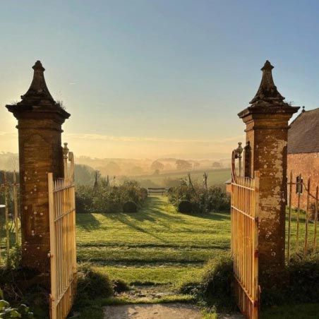 Alt text for Bettiscombe: "Looking through twin brick gateposts with decorative stone tops, framing a wooden gate, the view opens onto the sweeping gardens of Bettiscombe Manor. Rolling green lawns stretch into the distance, lined with mature trees and flowering borders, with the Dorset countryside and Vale beyond. The early morning or late afternoon light casts long shadows across the grass, and a soft mist hangs gently over the distant landscape beneath a pale blue sky. This is the entrance to designer Jasper Conran's Bettiscombe Manor near Bridport, which opens for the National Garden Scheme on Thursday 11 June. The gardens showcase Conran's creative, constantly evolving planting with orchards, mellow brick enclosures, and broad beds flanking an unforgettable view down to the sea