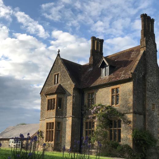 Pugin Hall, a Grade I listed Victorian stone building near Dorchester, stands majestically against a blue sky with white clouds. The honey-coloured stone house features distinctive Gothic-style windows, tall twin chimneys, and climbing ivy covering parts of the walls. The building is set within expansive green parkland on a gently sloping site.  It opens for the National Garden Scheme on Sunday 14 June