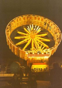 The Round-Up ride at Portland Fair glowing against the night sky, its great tilted wheel lit up in golden yellow lights with illuminated spokes radiating from the centre. The spinning drum is caught mid-motion, blurring the lights around its rim, and a sunburst design shines at its base above the pay booth, where hand-painted lettering reads The Sensational Round-Up. Silhouetted figures of fairgoers stand watching in the darkness below, with the metal barriers of the ride just visible in the foreground. The photograph captures the dazzling spectacle of the fair after dark, when Chiswell filled with light, noise and crowds every November.
