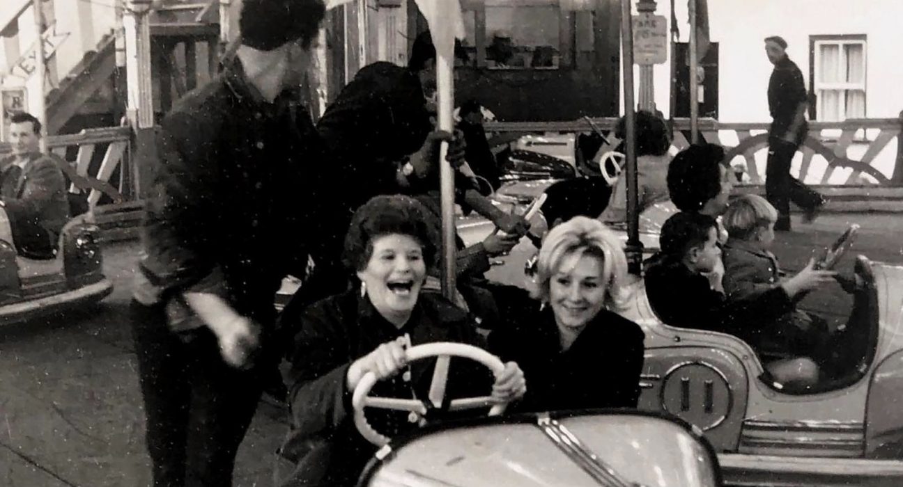 A black and white archive photograph of Portland Fair showing two women laughing on the dodgems. The woman driving has dark curled hair and dangling earrings, her mouth open mid-laugh as she grips the white steering wheel, while her blonde companion smiles beside her. Behind them, a fairground worker in a dark shirt steps between the bumper cars, and children can be seen riding in other cars across the rink. A man walks past the painted boards at the edge of the ride, with strings of lights and fairground signage visible above. The photograph captures the joy and atmosphere of the much-missed fair, held in Chiswell every November 5 for more than 200 years until it ended in 2001.