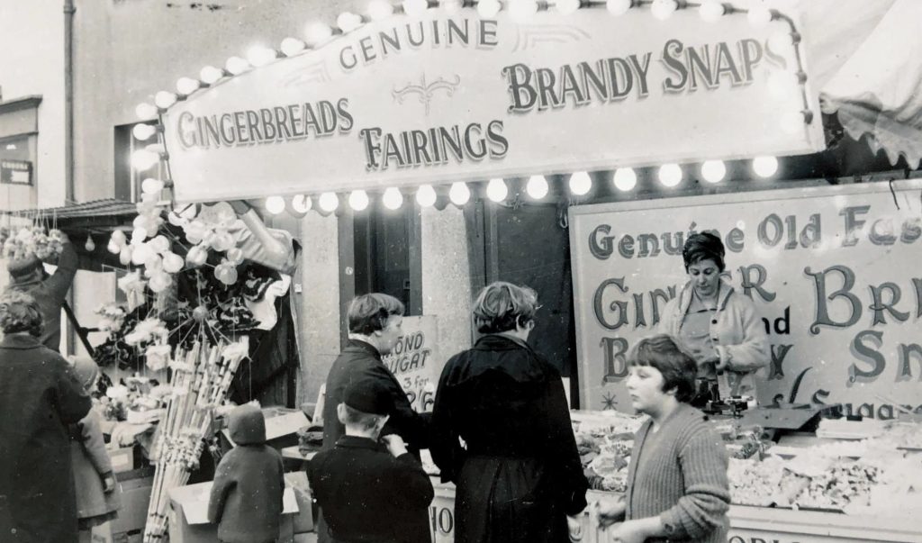A black and white archive photograph of a sweet stall at Portland Fair, its hand-painted sign lit by rows of bulbs and advertising genuine gingerbreads, fairings and brandy snaps. A woman in a light cardigan serves behind the counter, which is piled with bags of sweets, while women in dark coats and children gather in front of the stall. To the left hang balloons and bundles of fairground treats, with a sign offering almond nougat three for half a crown. A young girl in a knitted jumper stands in the foreground, and the buildings of Chiswell can be glimpsed behind the stall's awning. The photograph captures the traditional fairground food stalls that drew islanders to the fair each November.