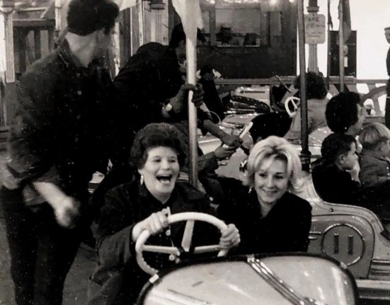 A black and white archive photograph of Portland Fair showing two women laughing on the dodgems. The woman driving has dark curled hair and dangling earrings, her mouth open mid-laugh as she grips the white steering wheel, while her blonde companion smiles beside her. Behind them, a fairground worker in a dark shirt steps between the bumper cars, and children can be seen riding in other cars across the rink. A man walks past the painted boards at the edge of the ride, with strings of lights and fairground signage visible above. The photograph captures the joy and atmosphere of the much-missed fair, held in Chiswell every November 5 for more than 200 years until it ended in 2001.