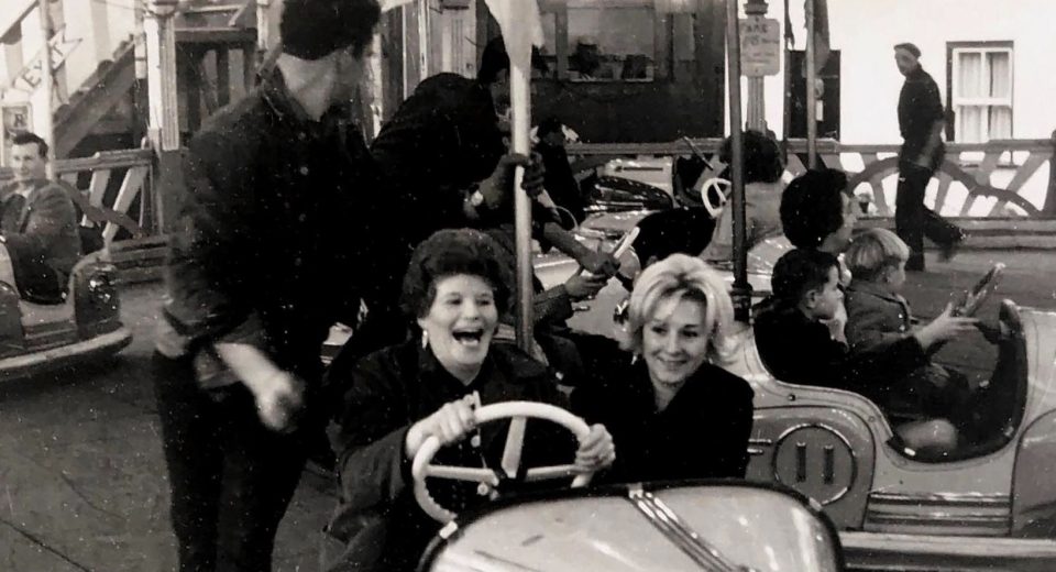 A black and white archive photograph of Portland Fair showing two women laughing on the dodgems. The woman driving has dark curled hair and dangling earrings, her mouth open mid-laugh as she grips the white steering wheel, while her blonde companion smiles beside her. Behind them, a fairground worker in a dark shirt steps between the bumper cars, and children can be seen riding in other cars across the rink. A man walks past the painted boards at the edge of the ride, with strings of lights and fairground signage visible above. The photograph captures the joy and atmosphere of the much-missed fair, held in Chiswell every November 5 for more than 200 years until it ended in 2001.