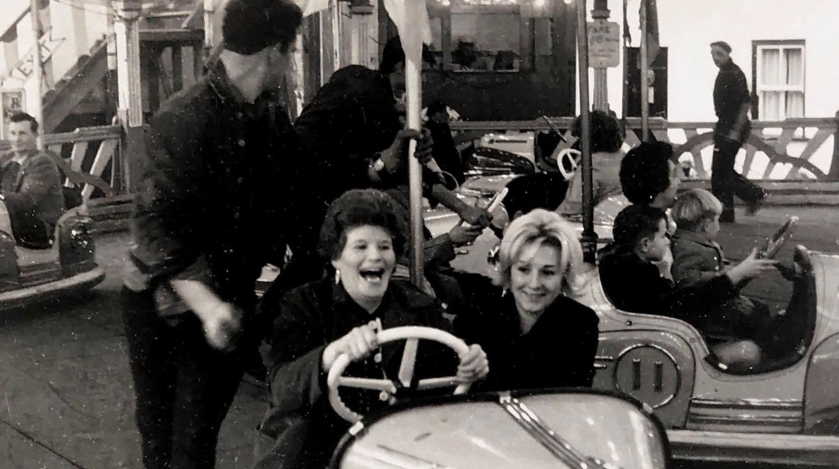 A black and white archive photograph of Portland Fair showing two women laughing on the dodgems. The woman driving has dark curled hair and dangling earrings, her mouth open mid-laugh as she grips the white steering wheel, while her blonde companion smiles beside her. Behind them, a fairground worker in a dark shirt steps between the bumper cars, and children can be seen riding in other cars across the rink. A man walks past the painted boards at the edge of the ride, with strings of lights and fairground signage visible above. The photograph captures the joy and atmosphere of the much-missed fair, held in Chiswell every November 5 for more than 200 years until it ended in 2001.