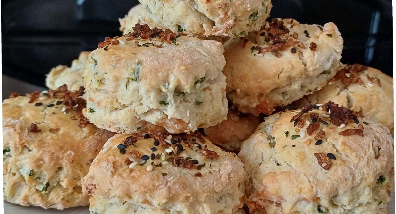 A pile of freshly baked smoked salmon and samphire scones stacked on a white plate, made using this savoury scones recipe. The scones are golden-brown on top, scattered with black and white sesame seeds and crispy onion pieces, with flecks of green samphire and pale pink salmon visible through the crumbly dough. The savoury scones are well risen with rough, craggy edges, and the plate sits on a dark kitchen worktop. The photograph shows the finished savoury scones recipe ready to be served, perfect for a summer picnic with a generous spread of cream cheese.