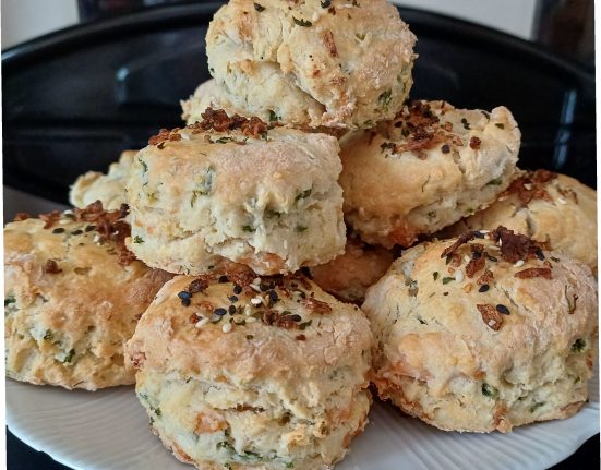 A pile of freshly baked smoked salmon and samphire scones stacked on a white plate, made using this savoury scones recipe. The scones are golden-brown on top, scattered with black and white sesame seeds and crispy onion pieces, with flecks of green samphire and pale pink salmon visible through the crumbly dough. The savoury scones are well risen with rough, craggy edges, and the plate sits on a dark kitchen worktop. The photograph shows the finished savoury scones recipe ready to be served, perfect for a summer picnic with a generous spread of cream cheese.