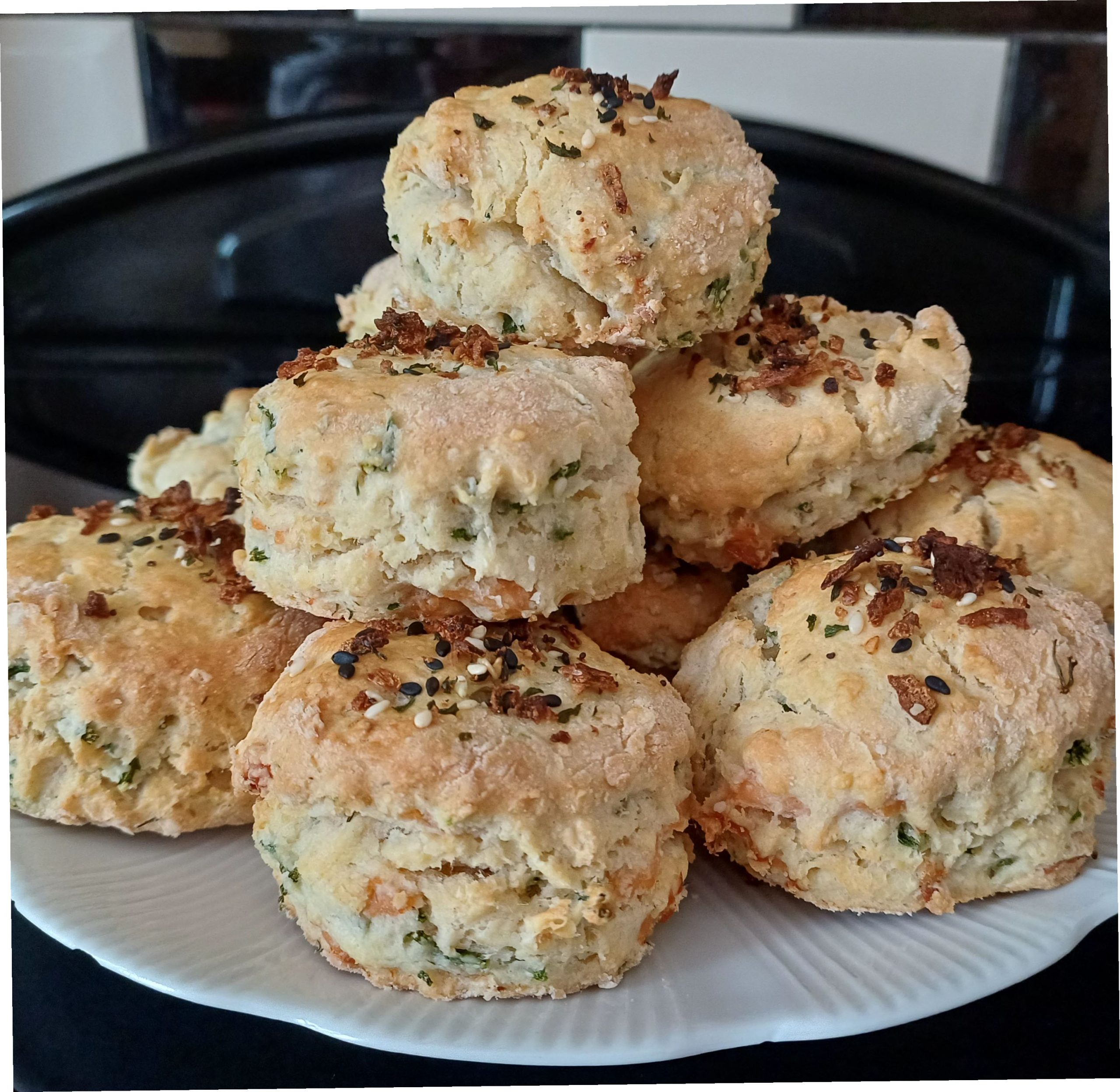 A pile of freshly baked smoked salmon and samphire scones stacked on a white plate, made using this savoury scones recipe. The scones are golden-brown on top, scattered with black and white sesame seeds and crispy onion pieces, with flecks of green samphire and pale pink salmon visible through the crumbly dough. The savoury scones are well risen with rough, craggy edges, and the plate sits on a dark kitchen worktop. The photograph shows the finished savoury scones recipe ready to be served, perfect for a summer picnic with a generous spread of cream cheese.