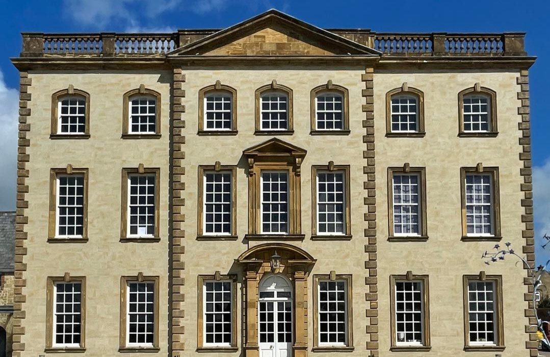 Sherborne House, a grand three-storey Georgian mansion built from golden hamstone, photographed front-on under a bright blue sky with wispy white clouds. The symmetrical facade features three rows of tall white sash windows with small square panes, each framed in honey-coloured stone surrounds. A central white double door sits beneath an ornate curved stone canopy with a lantern hanging inside it, and directly above is a striking full-height arched window with a carved triangular pediment. The roofline is crowned with a stone balustrade and a central pediment. A pale stone path leads to the entrance between two neat lawns, with small clipped shrubs lining the front of the building and a sculptural metal bird artwork just visible to the right