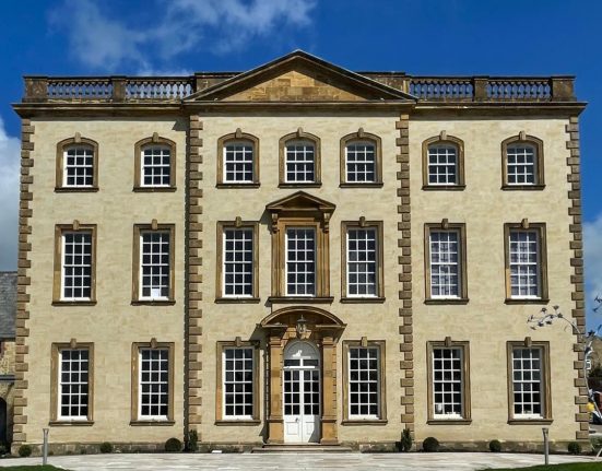 Sherborne House, a grand three-storey Georgian mansion built from golden hamstone, photographed front-on under a bright blue sky with wispy white clouds. The symmetrical facade features three rows of tall white sash windows with small square panes, each framed in honey-coloured stone surrounds. A central white double door sits beneath an ornate curved stone canopy with a lantern hanging inside it, and directly above is a striking full-height arched window with a carved triangular pediment. The roofline is crowned with a stone balustrade and a central pediment. A pale stone path leads to the entrance between two neat lawns, with small clipped shrubs lining the front of the building and a sculptural metal bird artwork just visible to the right