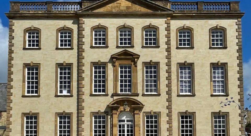 Sherborne House, a grand three-storey Georgian mansion built from golden hamstone, photographed front-on under a bright blue sky with wispy white clouds. The symmetrical facade features three rows of tall white sash windows with small square panes, each framed in honey-coloured stone surrounds. A central white double door sits beneath an ornate curved stone canopy with a lantern hanging inside it, and directly above is a striking full-height arched window with a carved triangular pediment. The roofline is crowned with a stone balustrade and a central pediment. A pale stone path leads to the entrance between two neat lawns, with small clipped shrubs lining the front of the building and a sculptural metal bird artwork just visible to the right
