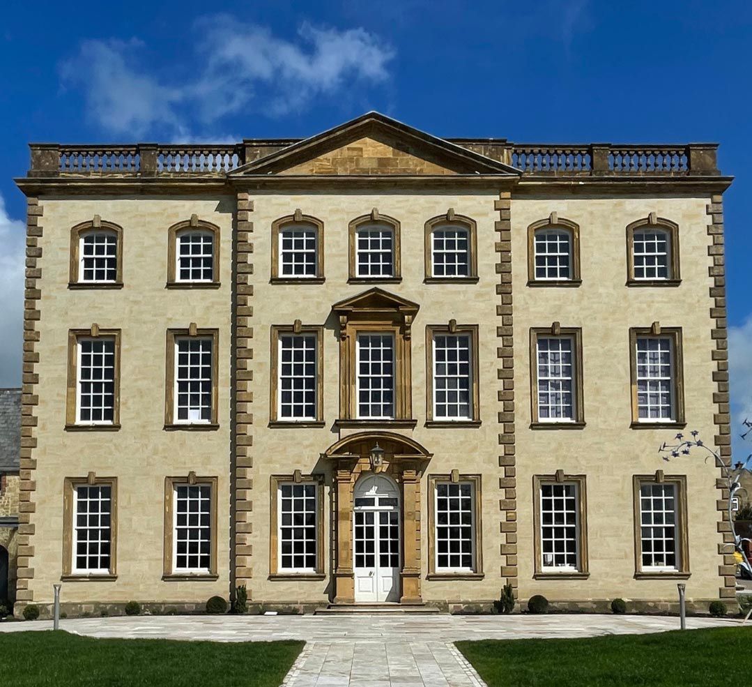 Sherborne House, a grand three-storey Georgian mansion built from golden hamstone, photographed front-on under a bright blue sky with wispy white clouds. The symmetrical facade features three rows of tall white sash windows with small square panes, each framed in honey-coloured stone surrounds. A central white double door sits beneath an ornate curved stone canopy with a lantern hanging inside it, and directly above is a striking full-height arched window with a carved triangular pediment. The roofline is crowned with a stone balustrade and a central pediment. A pale stone path leads to the entrance between two neat lawns, with small clipped shrubs lining the front of the building and a sculptural metal bird artwork just visible to the right