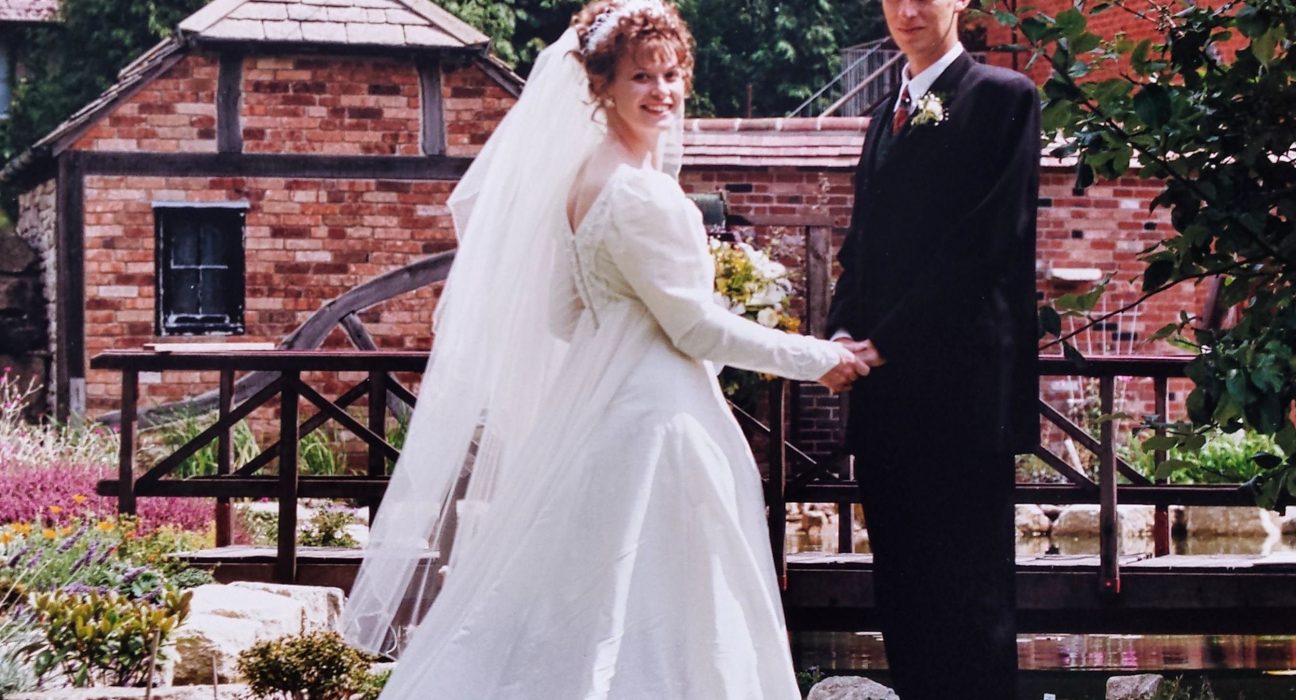 Liz, in a white wedding dress and long veil, holds the hand of her groom Phil in a dark suit and burgundy tie as they pose together in the manicured gardens of Springfield Country Hotel on their wedding day in 1996. Behind them stands the historic red-brick hotel building with ivy covering its walls, surrounded by flowering shrubs, ornamental plants and lush green lawns. The couple faces the camera with Phil standing behind Liz, creating a classic wedding portrait set against the hotel's elegant country backdrop.
