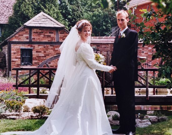 Liz, in a white wedding dress and long veil, holds the hand of her groom Phil in a dark suit and burgundy tie as they pose together in the manicured gardens of Springfield Country Hotel on their wedding day in 1996. Behind them stands the historic red-brick hotel building with ivy covering its walls, surrounded by flowering shrubs, ornamental plants and lush green lawns. The couple faces the camera with Phil standing behind Liz, creating a classic wedding portrait set against the hotel's elegant country backdrop.