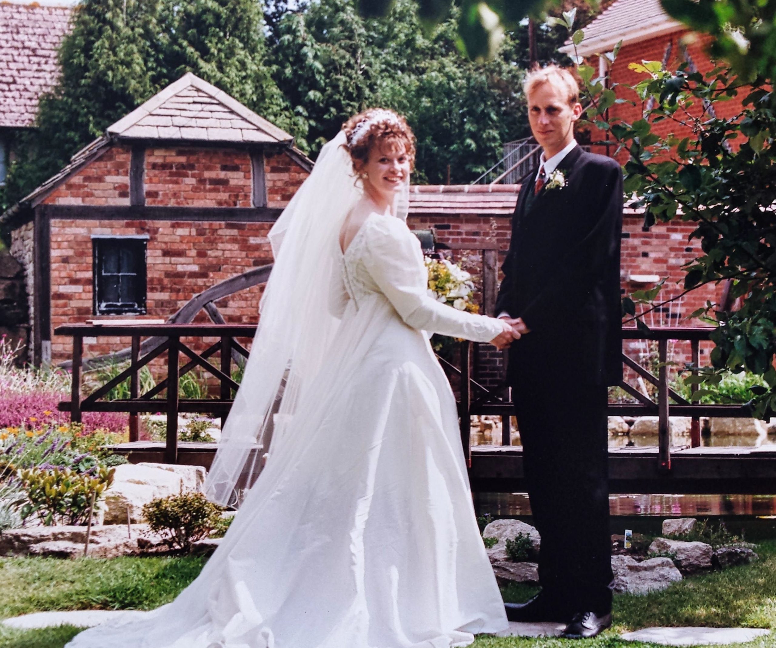 Liz, in a white wedding dress and long veil, holds the hand of her groom Phil in a dark suit and burgundy tie as they pose together in the manicured gardens of Springfield Country Hotel on their wedding day in 1996. Behind them stands the historic red-brick hotel building with ivy covering its walls, surrounded by flowering shrubs, ornamental plants and lush green lawns. The couple faces the camera with Phil standing behind Liz, creating a classic wedding portrait set against the hotel's elegant country backdrop.