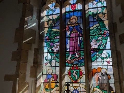 A stained glass window in St Giles Church, Hooke, featuring multiple panels with religious imagery rendered in vibrant colors including blues, greens, reds, yellows, and purples. The ornate window depicts various figures and scenes arranged vertically within a traditional lead framework and arched top. Sunlight streams through the colored glass, illuminating the intricate details. A black metal railing stands in front of the window.