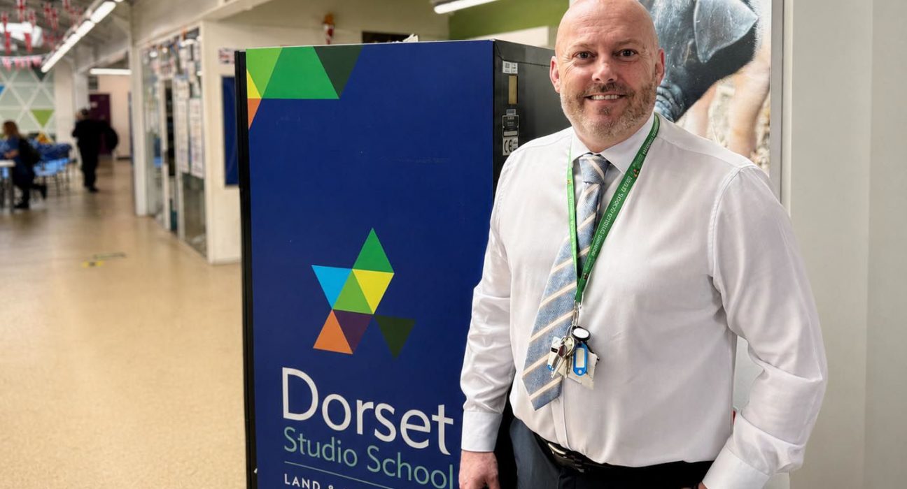 Jason Malone, Principal of Dorset Studio School, smiles at the camera while standing in a bright school corridor next to the school's branded signage. The blue sign features Dorset Studio School's distinctive geometric triangle logo in green, blue, yellow and orange, with "LAND & ENVIRO" visible below the school name. Jason wears a white shirt, patterned tie and a green lanyard with keys and ID badge. The modern school corridor stretches behind him with fluorescent lighting and students visible in the background.