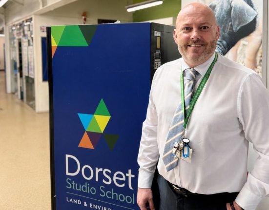 Jason Malone, Principal of Dorset Studio School, smiles at the camera while standing in a bright school corridor next to the school's branded signage. The blue sign features Dorset Studio School's distinctive geometric triangle logo in green, blue, yellow and orange, with "LAND & ENVIRO" visible below the school name. Jason wears a white shirt, patterned tie and a green lanyard with keys and ID badge. The modern school corridor stretches behind him with fluorescent lighting and students visible in the background.