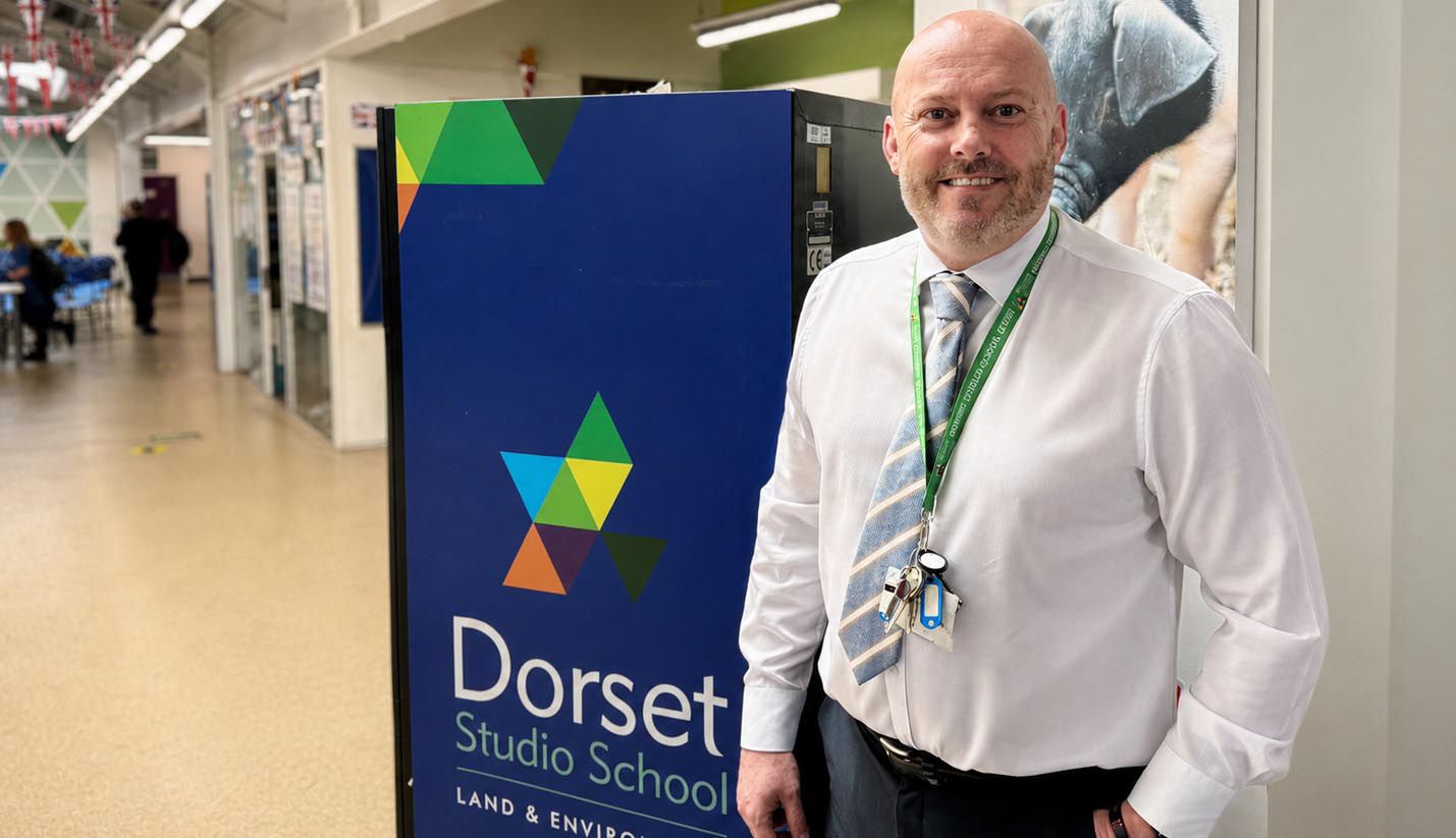 Jason Malone, Principal of Dorset Studio School, smiles at the camera while standing in a bright school corridor next to the school's branded signage. The blue sign features Dorset Studio School's distinctive geometric triangle logo in green, blue, yellow and orange, with "LAND & ENVIRO" visible below the school name. Jason wears a white shirt, patterned tie and a green lanyard with keys and ID badge. The modern school corridor stretches behind him with fluorescent lighting and students visible in the background.