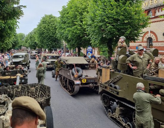 A WWII military heritage parade on a tree-lined town street. Multiple vintage military vehicles including Jeeps, trucks and tanks are positioned on the road, crewed by people in authentic WWII-era military uniforms. Large crowds of spectators line both sides of the street beneath mature green trees. A brick building with "Brewery Square" visible can be seen on the right side. British flags and period-appropriate signage are visible among the crowds. The scene captures a public commemoration or heritage event celebrating Second World War military history.