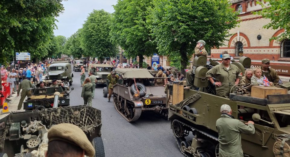 A WWII military heritage parade on a tree-lined town street. Multiple vintage military vehicles including Jeeps, trucks and tanks are positioned on the road, crewed by people in authentic WWII-era military uniforms. Large crowds of spectators line both sides of the street beneath mature green trees. A brick building with "Brewery Square" visible can be seen on the right side. British flags and period-appropriate signage are visible among the crowds. The scene captures a public commemoration or heritage event celebrating Second World War military history.