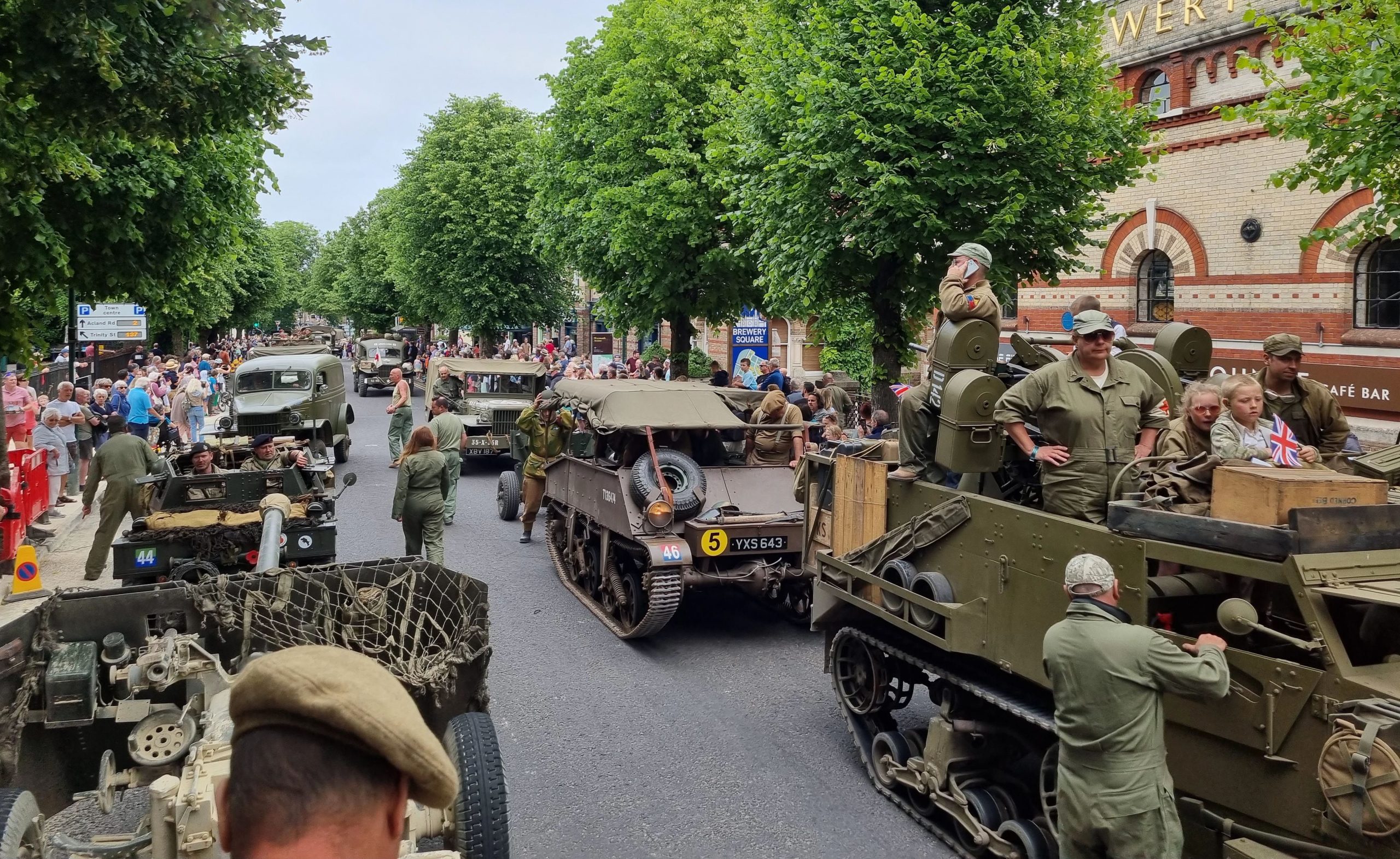 A WWII military heritage parade on a tree-lined town street. Multiple vintage military vehicles including Jeeps, trucks and tanks are positioned on the road, crewed by people in authentic WWII-era military uniforms. Large crowds of spectators line both sides of the street beneath mature green trees. A brick building with "Brewery Square" visible can be seen on the right side. British flags and period-appropriate signage are visible among the crowds. The scene captures a public commemoration or heritage event celebrating Second World War military history.