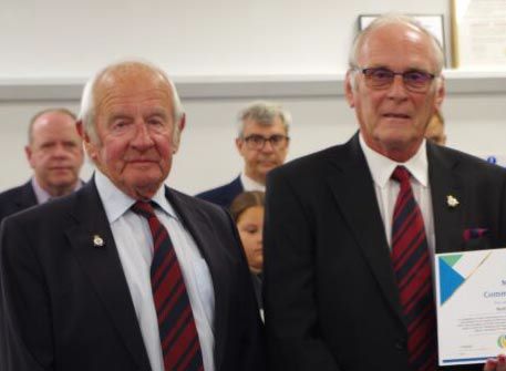 Two older men in dark suits with red ties, holding a blue and white Community Award certificate, at Weymouth's 2026 Mayor's Community Awards ceremony. They are Nothe Fort volunteers being recognised for their dedication to maintaining the historic fort and hosting events for the community