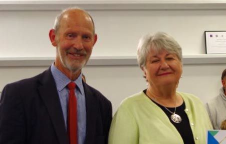 Richard Sloan, wearing a blue and white striped shirt and red tie with a reddish beard, stands beside Liz Bartlett, who wears a bright yellow-green cardigan over a black top with a decorative brooch and has short white hair. Both are smiling at Weymouth's 2026 Mayor's Community Awards ceremony, where they were recognised for their dedication and commitment to St Ann's and St Aldhelm's churches. Framed certificates are visible on shelves behind them.