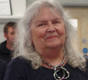 Sue Prescott, a woman with long white hair and a thoughtful expression, wears a dark navy top and an eye-catching multi-coloured beaded necklace with interlocking rings of blue, green, purple and other colours. She is pictured at Weymouth's 2026 Mayor's Community Awards ceremony, where she was recognised for her decades of dedicated community work across Weymouth. In the background, partially out of focus, another person can be seen. Shelving with notices is visible behind her