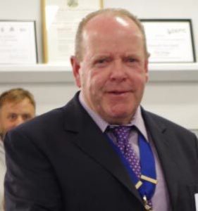 Dave Lane, chairman of Weymouth Angling Society, stands at Weymouth's 2026 Mayor's Community Awards ceremony. He is a man with reddish-blonde hair, balding on top, with a warm smile. He wears a dark suit jacket and a purple and blue patterned tie, with a medal or pin on his chest. Behind him, framed certificates are visible on shelving. Dave accepted the award on behalf of Weymouth Angling Society in recognition of over 125 years of encouraging and supporting line fishing for all age groups