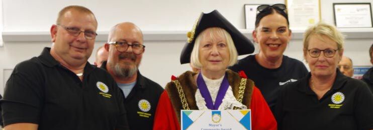 he Mayor, wearing ceremonial robes with red and gold trim and a purple ribbon, stands in the centre holding a Community Award certificate, flanked by four volunteers from Secret Behind The Smile. On the left, a man wearing glasses and a dark polo shirt with the organisation's logo smiles warmly next to another man also in a dark polo shirt with glasses. On the right, a woman in a dark polo shirt with a Nike logo and another woman wearing glasses in a dark polo shirt with the organisation's logo complete the group. All are beaming at Weymouth's 2026 Mayor's Community Awards ceremony. Framed certificates are visible on shelves behind them. Secret Behind The Smile was recognised for their volunteers' dedication to supporting people living with stoma and helping rebuild body confidence in those adjusting to life with an ostomy.