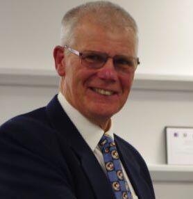 Alan Hale, a man with reddish-grey hair and glasses, beams with a warm smile at Weymouth's 2026 Mayor's Community Awards ceremony. He wears a dark suit jacket and a patterned tie with a medal or pin on his chest, indicating his long service. Behind him, shelving with framed certificates is visible. Alan was recognised for over 55 years of dedicated voluntary commitment to the Royal Life Saving Society, delivering free defibrillator and community CPR training sessions across Weymouth and the surrounding area, helping to make the community a safer place