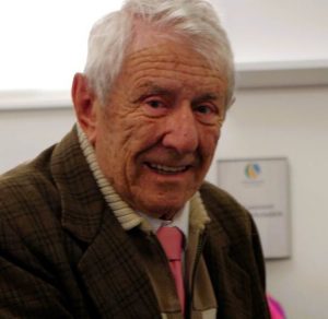 Eric Coetzee, Weymouth Museum's longest-serving volunteer, beams with genuine warmth and joy at Weymouth's 2026 Mayor's Community Awards ceremony. He is an older man with thick white hair, his face lined with age and experience, eyes twinkling with a bright smile. He wears an olive-brown jacket over a white shirt with a pink and coral tie. Behind him, a chart or notice board is visible on the wall. Eric was recognised in recognition of his more than 30 years of dedicated service to Weymouth Museum. His contribution, commitment and professionalism is highly valued by the museum