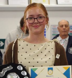 Bethany-Mae Paice (Beth), a young woman with reddish-blonde hair and glasses with dark frames, smiles confidently while holding her Mayor's Community Award at Weymouth's 2026 Mayor's Community Awards ceremony. She wears a cream coloured top with black polka dots and a warm brown cardigan, looking proud and pleased. In the background, other attendees and shelving with notices are visible. Beth was recognised as Youth Councillor of the Year for her role as Chair of Weymouth's Youth Council, representing young people of the town and supporting other members in numerous events and community projects they have initiated