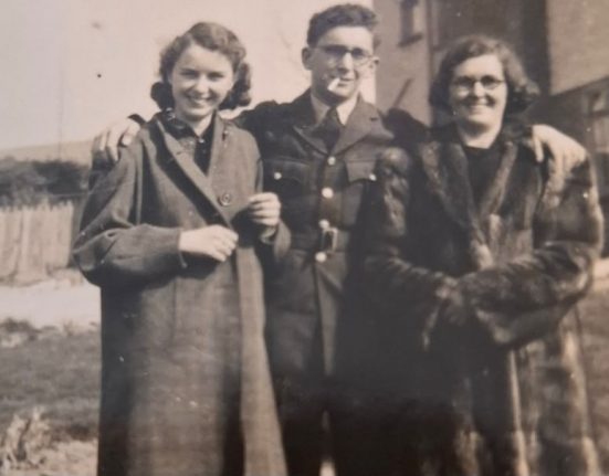 Black and white photograph from March 1941 showing three people standing together outdoors near the coast. A young woman stands on the left wearing a dark coat over a blouse, her hair styled in soft waves typical of the era. In the centre, a man in uniform, likely William Paul Tizzard, stands smartly dressed in military uniform with a cap, hands by his sides. To the right, another man in similar military uniform stands slightly behind, also wearing a cap. The group stands on a grassy verge with a low wall or embankment behind them, and a hazy view of the sea or coastline visible in the background, suggesting a wartime photograph taken near the Weymouth coast before deployment.