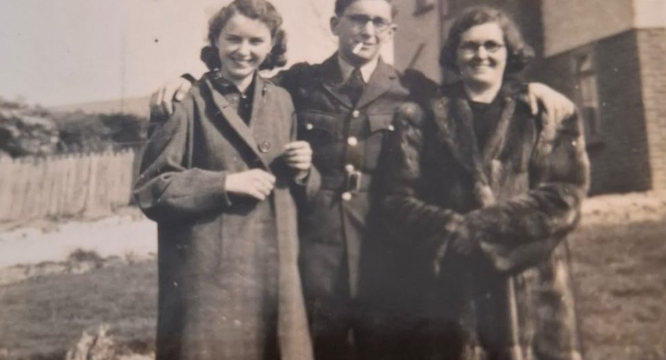 Black and white photograph from March 1941 showing three people standing together outdoors near the coast. A young woman stands on the left wearing a dark coat over a blouse, her hair styled in soft waves typical of the era. In the centre, a man in uniform, likely William Paul Tizzard, stands smartly dressed in military uniform with a cap, hands by his sides. To the right, another man in similar military uniform stands slightly behind, also wearing a cap. The group stands on a grassy verge with a low wall or embankment behind them, and a hazy view of the sea or coastline visible in the background, suggesting a wartime photograph taken near the Weymouth coast before deployment.