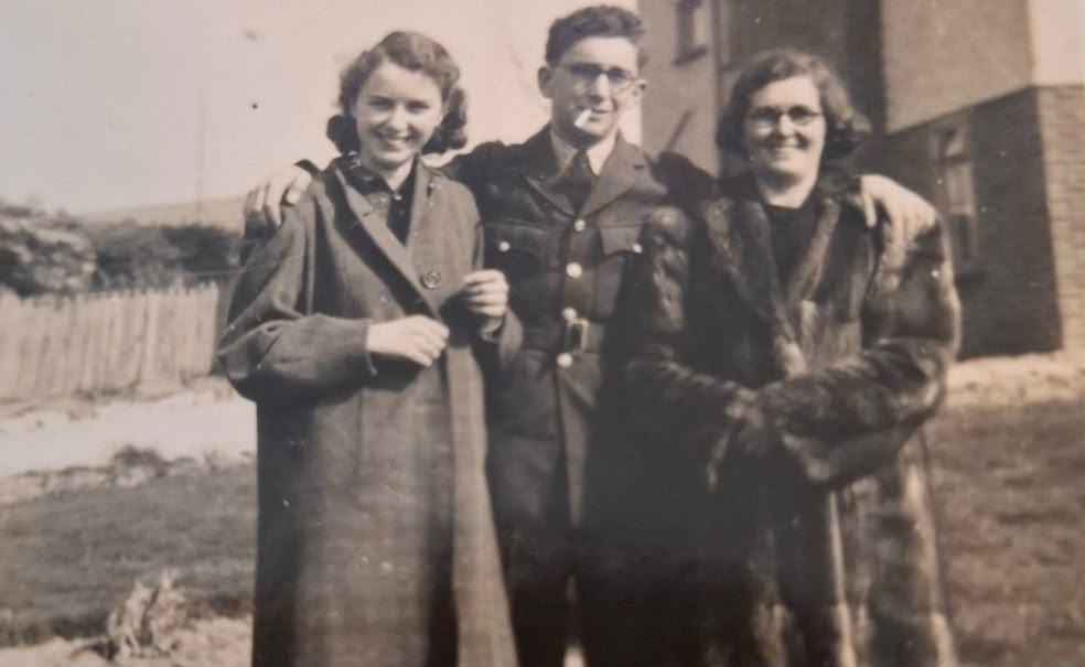 Black and white photograph from March 1941 showing three people standing together outdoors near the coast. A young woman stands on the left wearing a dark coat over a blouse, her hair styled in soft waves typical of the era. In the centre, a man in uniform, likely William Paul Tizzard, stands smartly dressed in military uniform with a cap, hands by his sides. To the right, another man in similar military uniform stands slightly behind, also wearing a cap. The group stands on a grassy verge with a low wall or embankment behind them, and a hazy view of the sea or coastline visible in the background, suggesting a wartime photograph taken near the Weymouth coast before deployment.