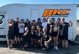 Group of BKC Racing drivers and team members celebrating with trophies in front of a BKC Racing van at Clay Pigeon Raceway.