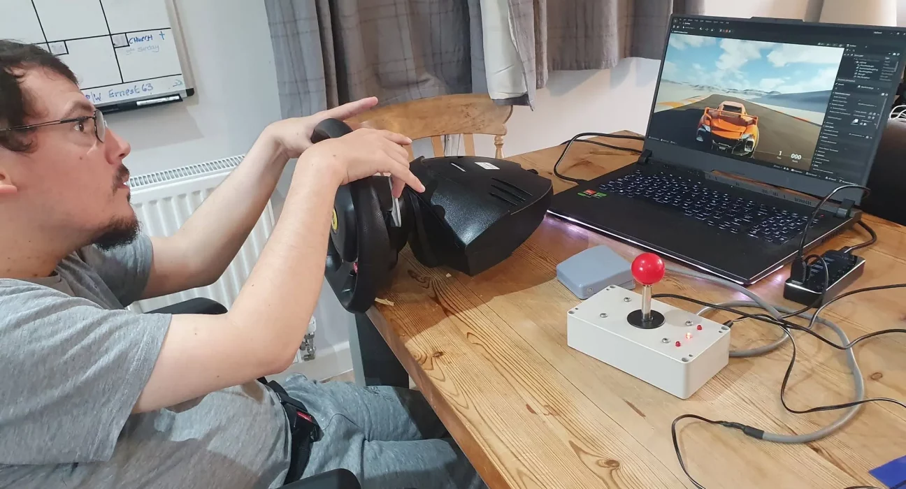 Louis Gale, founder of the Accessible Gaming Café in Dorchester, using an adapted steering wheel and joystick controller to play a racing game on his laptop.