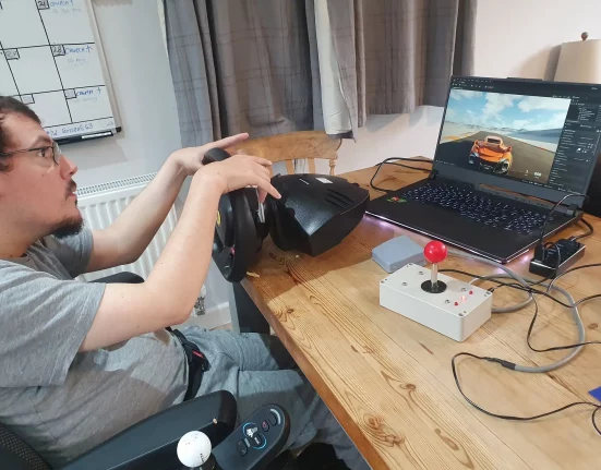 Louis Gale, founder of the Accessible Gaming Café in Dorchester, using an adapted steering wheel and joystick controller to play a racing game on his laptop.