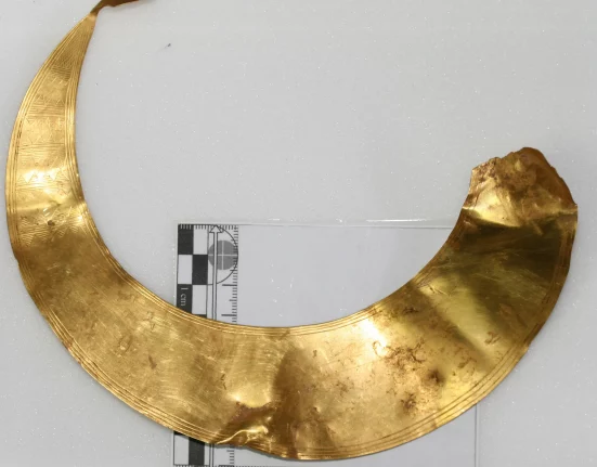 Bronze Age gold lunula found by metal detectors in Dorset's Tarrant Valley, a thin crescent-shaped neck ornament with geometric patterns, laid flat on a white background next to a centimetre scale ruler.