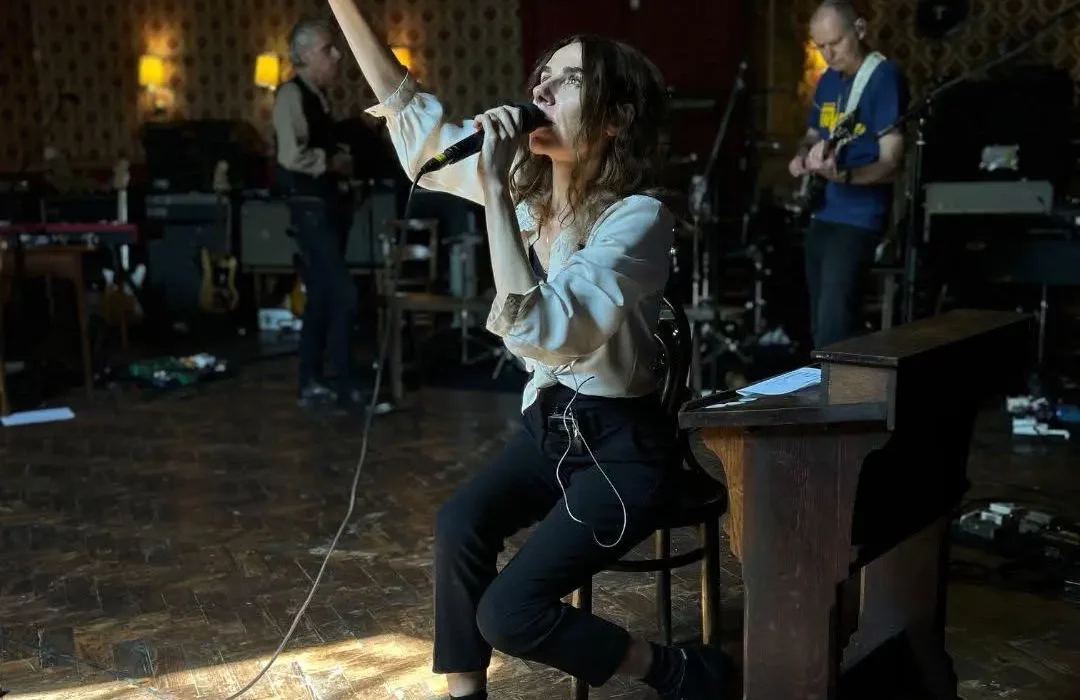 PJ Harvey sings with her arm raised while rehearsing with her band inside Bridport's Lyric Theatre, seated among red-and-gold flocked wallpaper and stage equipment.