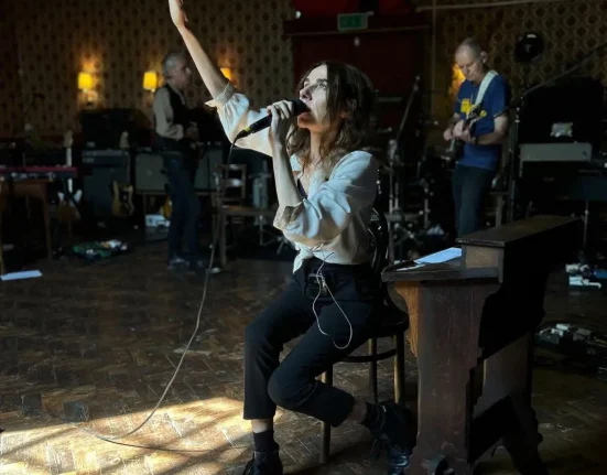 PJ Harvey sings with her arm raised while rehearsing with her band inside Bridport's Lyric Theatre, seated among red-and-gold flocked wallpaper and stage equipment.