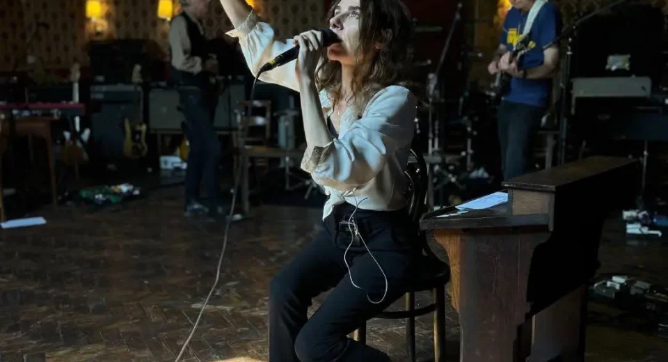 PJ Harvey sings with her arm raised while rehearsing with her band inside Bridport's Lyric Theatre, seated among red-and-gold flocked wallpaper and stage equipment.